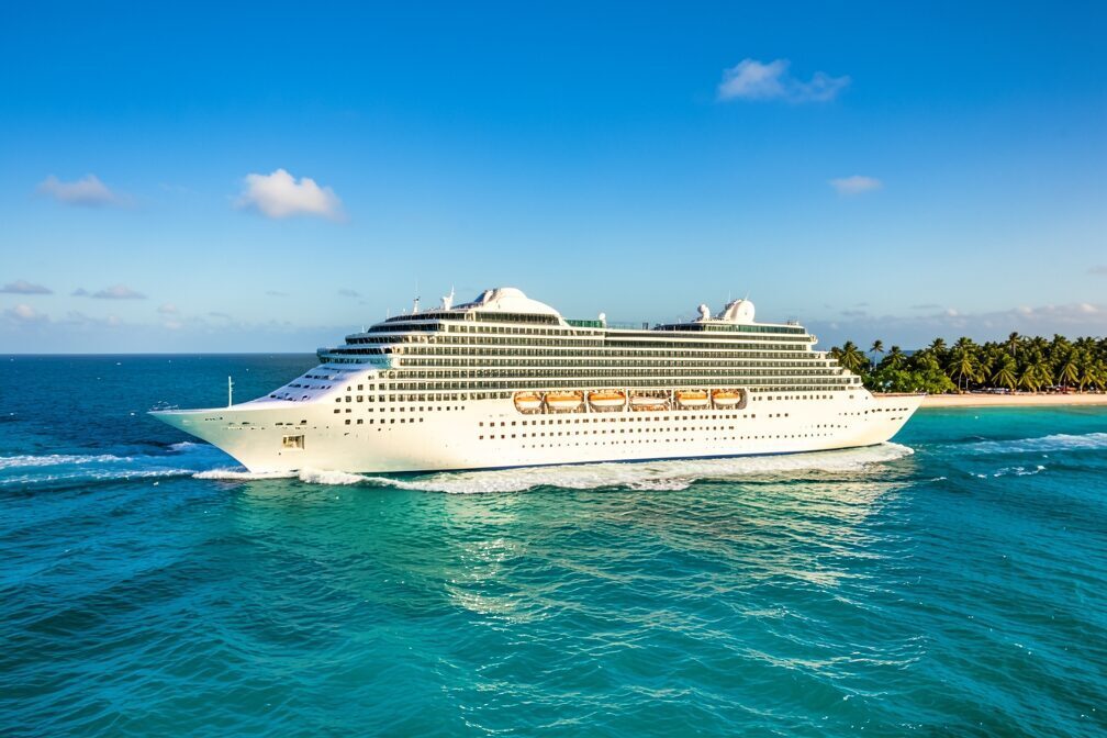 A large modern cruise ship sailing under full power across the brilliant turquoise water off Great Stirrup Cay in the Bahamas, white wake trailing behind, palm-fringed sand and a bright blue sky in the distance