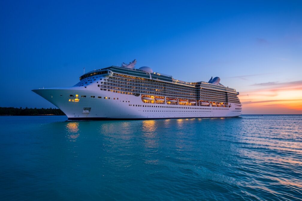 A large cruise ship sailing at dusk through the calm turquoise water off Great Stirrup Cay in the Bahamas, warm cabin lights glowing along its decks beneath a deep blue December sky